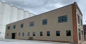 A two-story building with tan metal siding, multiple rectangular windows, and a brick corner sits next to large white silos under a cloudy sky—ideal for businesses specializing in crawlspace repair.