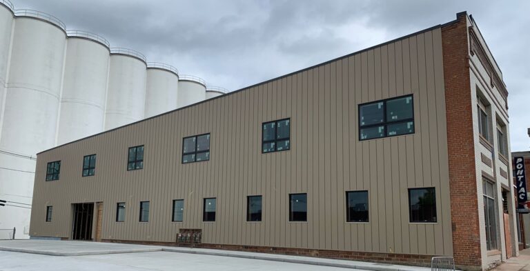 A two-story building with tan metal siding, multiple rectangular windows, and a brick corner sits next to large white silos under a cloudy sky—ideal for businesses specializing in crawlspace repair.