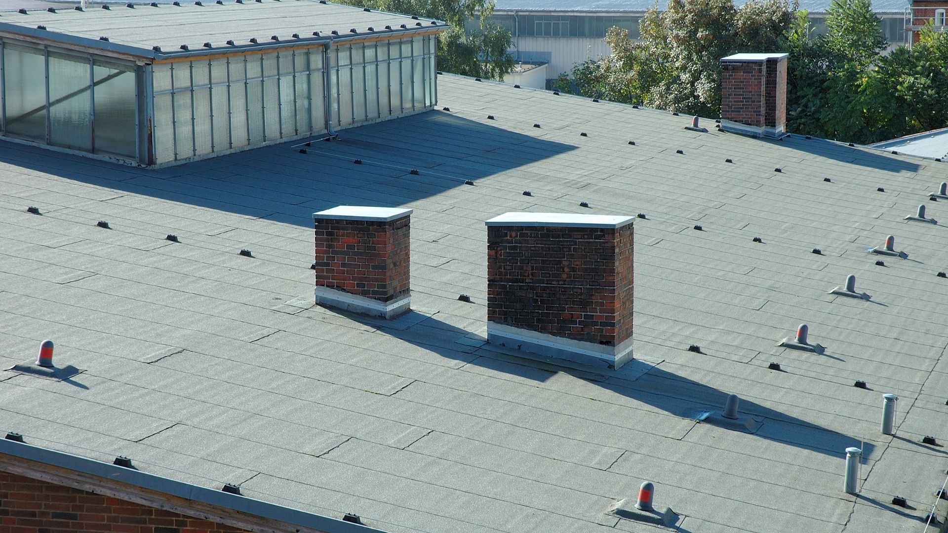 Flat industrial roof with two brick chimneys, multiple roof vents, and a section of glass windows near the top left; trees are visible in the background, highlighting a structure that may benefit from routine maintenance and foundation repair.