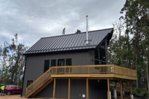 A modern, dark gray house with a metal roof, large windows, wood deck, and exterior staircase, surrounded by trees. A person and red car are visible on the left.