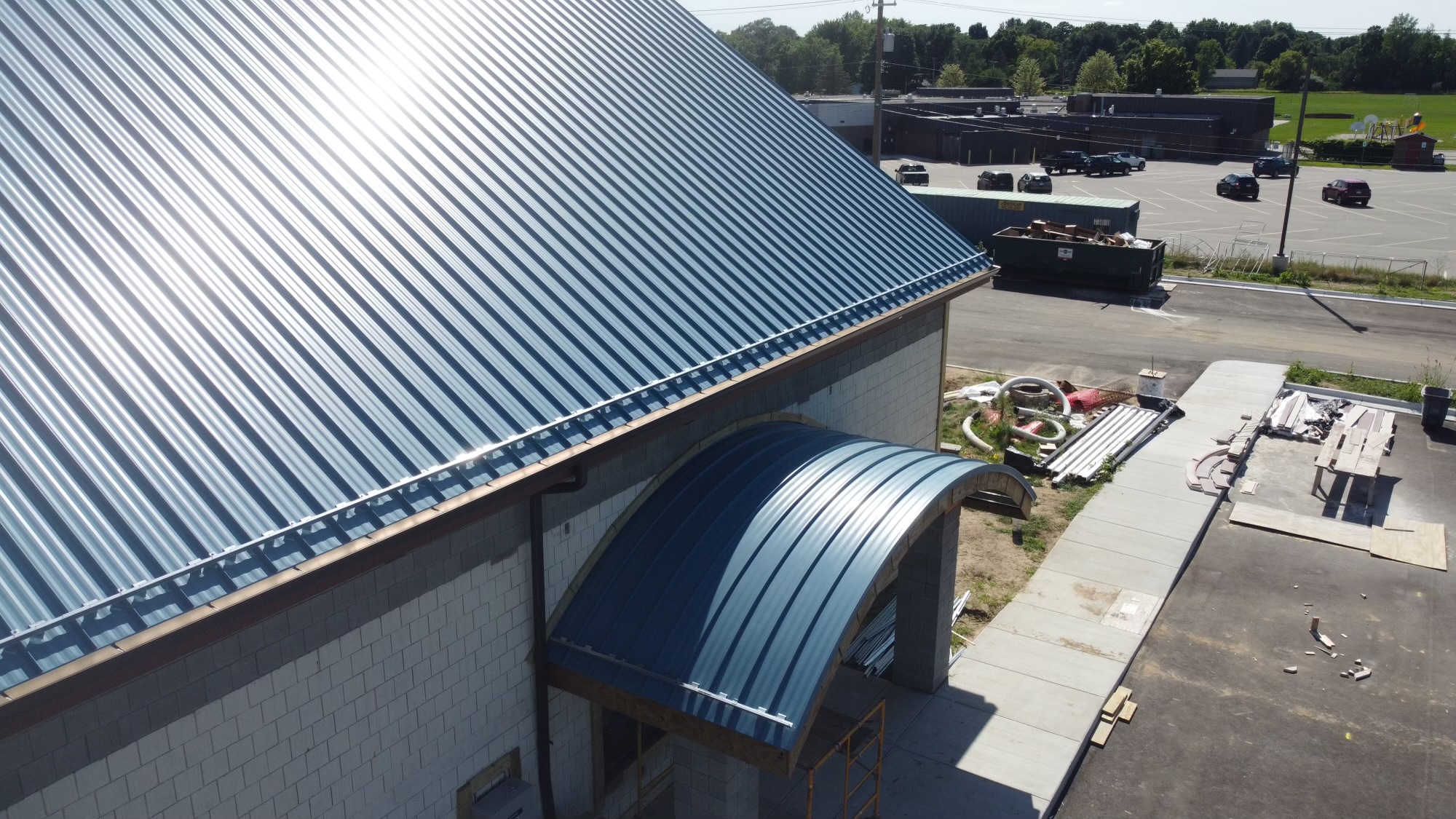 A building with a blue metal roof and awning under construction, with construction materials and vehicles visible in the adjacent parking lot.