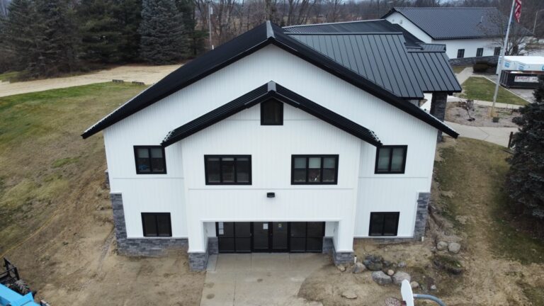 A two-story white building with black trim and a metal roof, recently updated with foundation repair, stands amid grass, rocks, and trees, viewed from above on a cloudy day.