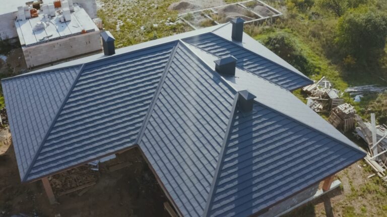 Aerial view of a modern house with a dark grey metal roof featuring multiple ridges and chimneys, surrounded by construction materials and vegetation—an ideal setting for observing ongoing foundation repair work.