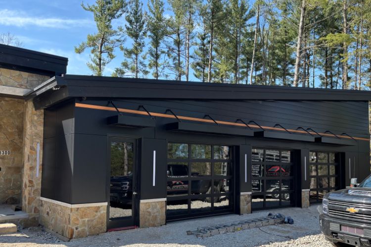 Modern black garage with glass doors and stone accents, surrounded by trees; a black pickup truck is parked in front on a gravel driveway.