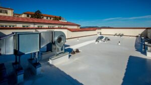 Flat rooftop with HVAC equipment and ventilation units, surrounded by a low wall. Foundation repair work may be underway, as nearby buildings with red-tiled roofs are visible in the background under a clear blue sky.