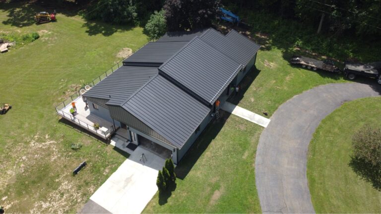 Aerial view of a modern house with a dark metal roof, concrete driveway, and surrounding green lawn, bordered by trees and a curved blacktop road.